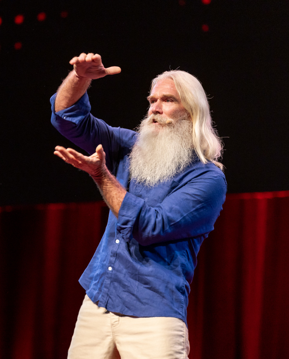 A man with a long white beard wearing a blue shirt stands on stage.